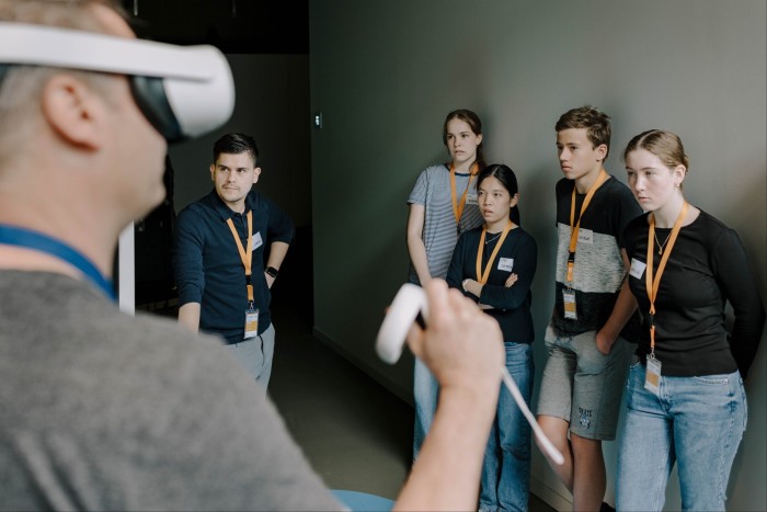 A group of young adults are standing attentively watching a man wearing a virtual reality headset