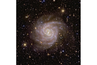 A pinkish and slightly hazy spiral galaxy in space, in front of many stars and more distant bright galaxies.