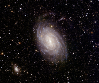 A beautiful spiral galaxy can be seen at the center of this image of space.  There are lots of stars in the background.  In the lower left corner of the scene, a slightly smaller hazy white spot.