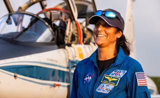 smiling female astronaut in flight suit and hat in front of jet plane with open cockpit