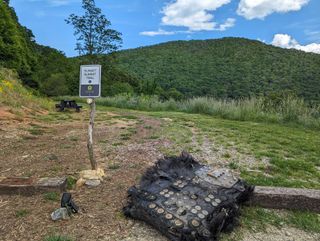 a large black piece of fiberglass covered in metal screws and plates lies on the ground next to a path leading into the forest.  rolling mountains can be seen in the distance