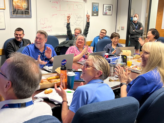 A bunch of people, many wearing blue, cheer in a conference room.