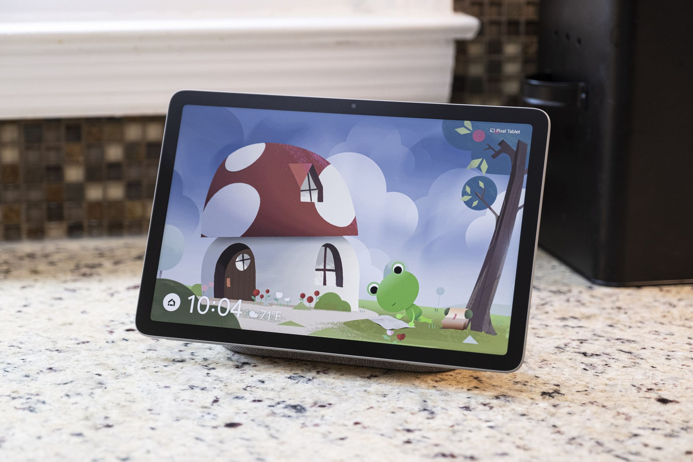 A Pixel Tablet mounted on a docking station with a speaker displaying the Weather Frog screensaver.