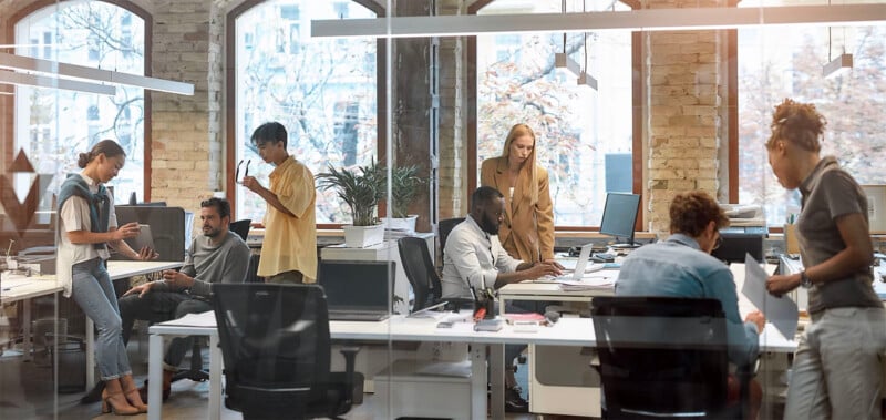 Modern office with brick walls and large windows.  Several people work at desks with computers while others stand and converse.  Plants and office equipment are spread around, creating a collaborative and bustling work environment.