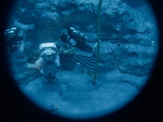 an astronaut in a space suit walks on the bottom of a pool next to a diver in a diving mask