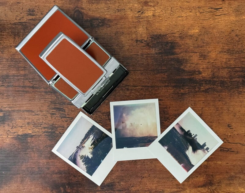 A vintage Polaroid camera with a brown and silver design sits on a wooden surface.  In front of the camera are three instant photos depicting outdoor scenes, including a forest, a body of water, and a cityscape with cloudy skies.