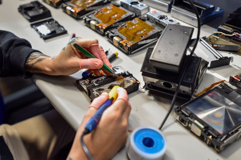 A person's hands are seen working at a table full of electronic components and tools.  One hand uses a soldering iron while the other holds a green tool.  Various disassembled devices and parts, including hard drives, are spread out on the desktop.