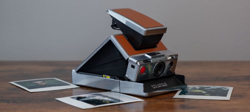 An old Polaroid SX-70 Land Camera with a brown leather exterior sits on a wooden surface.  The camera is partially unfolded.  Two instant photos are scattered in the foreground, partially visible.  The background is a plain gray wall.