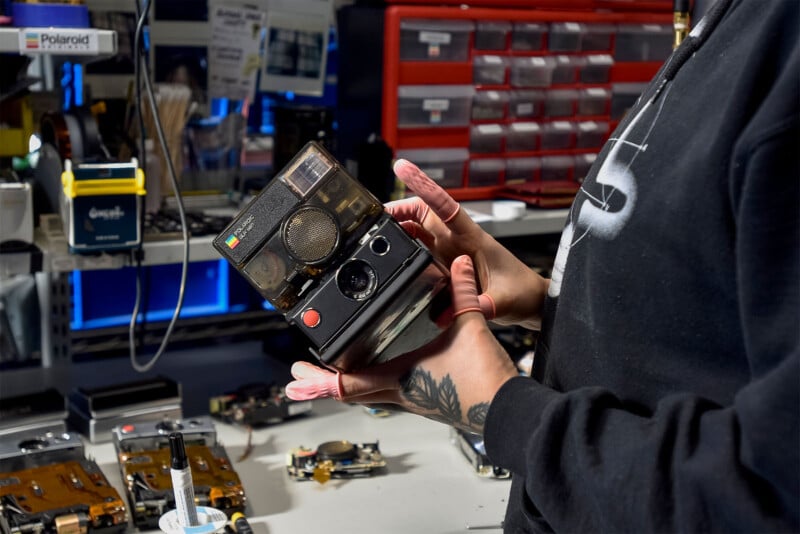 A person holding a vintage Polaroid camera inside a workshop.  The background contains various electronic components, tools and storage drawers to indicate a repair or restoration environment.  A tattoo on the person's hand and a partial view of their sweatshirt can be seen.