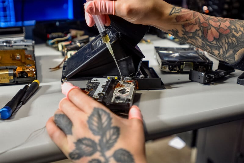 A person with tattooed hands and wearing pink gloves is repairing electronic components at a workstation.  They use a syringe to apply liquid to the part while various tools and electronic devices are laid out on the table.