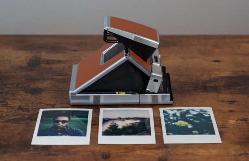 A vintage instant camera with a brown exterior sits on a wooden surface.  In front of her are three Polaroid photos: one of a person wearing sunglasses, one of a scenic view with a body of water, and one of yellow flowers in the foreground.