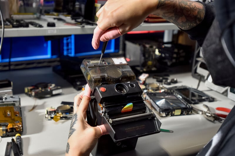 A person with tattooed arms uses a screwdriver to repair or assemble an old Polaroid camera at a workstation.  Various electronic components and tools are scattered on a white table around the workspace.