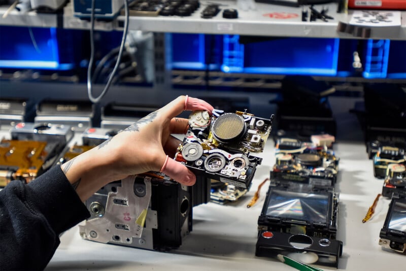 A person holds a disassembled mechanical part in a workshop, possibly from an old electronic device.  The work area is crowded with other disassembled parts and tools, in the background are shelves illuminated by blue lights.