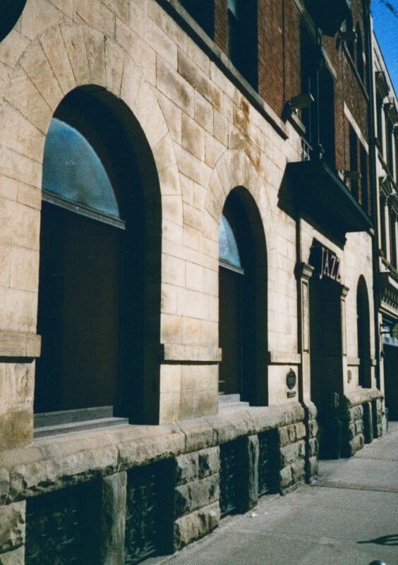 Street view of historic beige stone building with tall arched windows.  The building has signs "JAZZ" above the entrance.  The architectural style is characterized by a combination of stone and brick materials with ornamental details.  Shadows fall across the sidewalk.