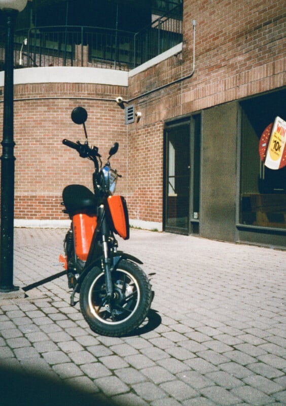 A red electric scooter stands on a cobbled street against a background of red brick buildings.  On the right, a yellow sign with a 70% discount is displayed in the shop window.  The area is brightly lit by the sun.