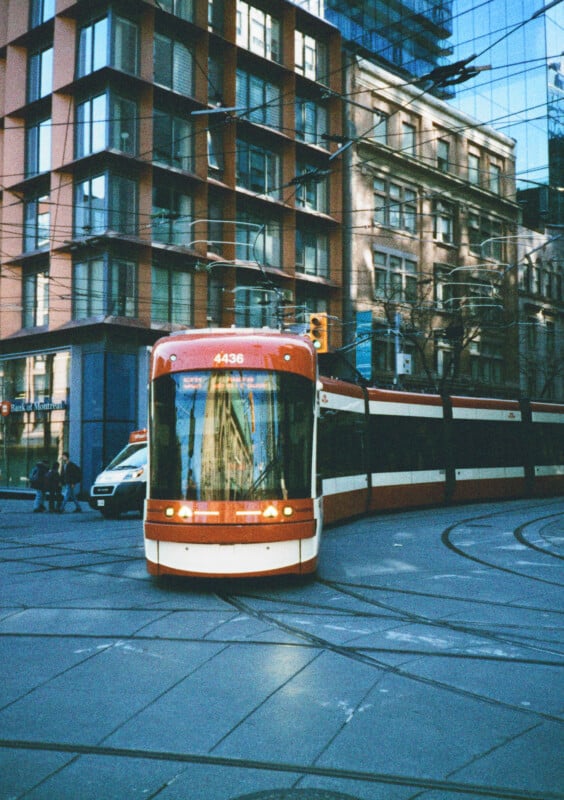 A red and white tram moves along the tracks on a city street corner with modern and older buildings in the background.  There is a van parked on the side of the street.  Reflections of buildings can be seen on the windows of the tram.