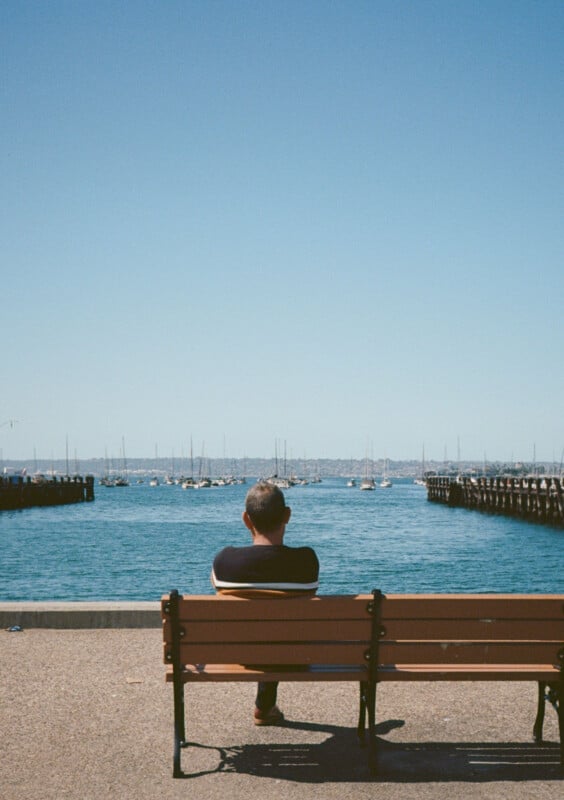 A man sits alone on a wooden bench, facing a body of water with many boats anchored in the distance.  The sky is clear and blue, stretching over the calm water and stretching to the horizon.  Two piers frame the scene on either side.