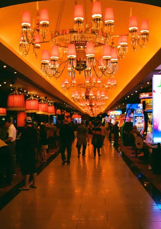 An interior scene of a vibrant casino features people walking and talking among slot machines and gaming tables.  The space is illuminated by ornate chandeliers and warm lighting, creating a lively atmosphere.  The ceiling is decorated with red lampshades.