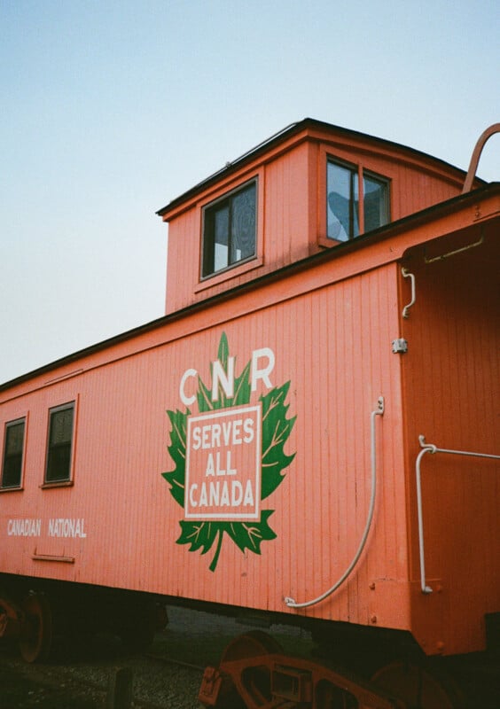 Canadian National Railway orange cab with "CNR serves all of Canada" written with a green leaf graphic on the side.  The cabin has a small cupola on the roof with a window and is placed outdoors with a clear sky in the background.