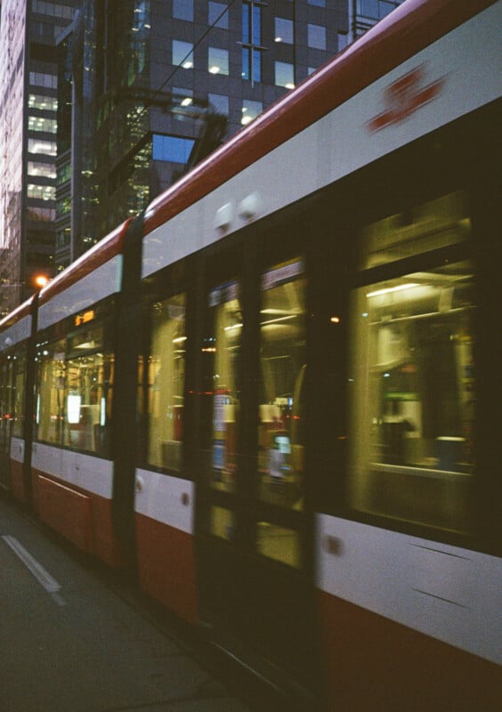 Motion blur photo of a modern light rail tram with red and white colors moving on a city street.  In the background are tall buildings with illuminated windows that reflect the city's evening scene.