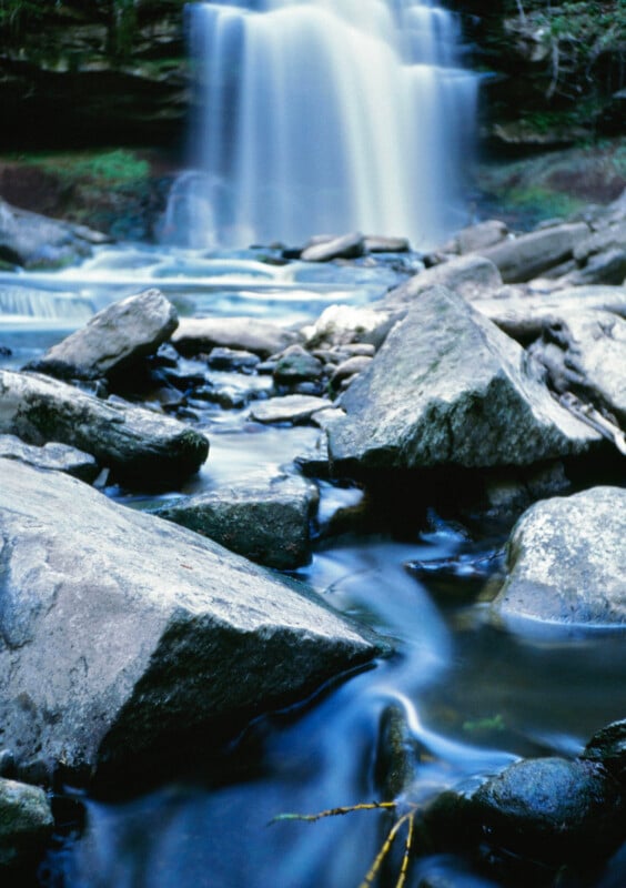 A tranquil waterfall tumbles over a cliff into a rocky stream.  Large stones and boulders are scattered in the water, creating a peaceful and natural scene.  Fresh greenery in the background enhances the calm atmosphere.