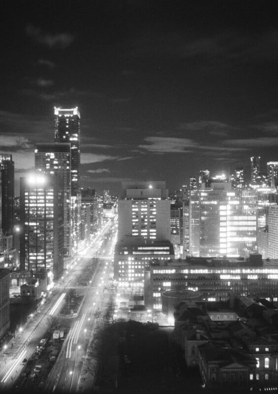 Aerial night view of the city, showing illuminated skyscrapers and buildings lining both sides of a wide, well-lit street with vehicle headlights spreading along it.  The sky is partly clear and the city lights create a glowing urban atmosphere.