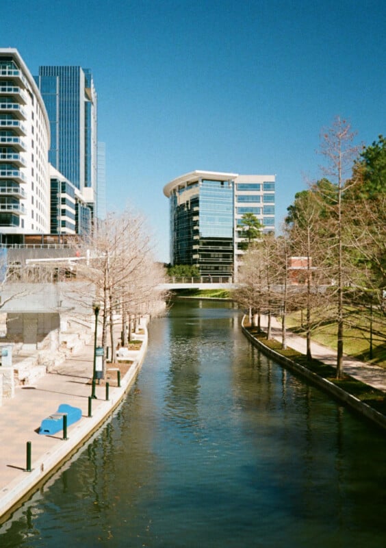 A peaceful city canal lined with bare trees on both sides, reflecting the clear blue sky.  Modern high rise buildings lining the waterway, with some greenery visible to the right.  A bridge in the distance connects the buildings over a canal.