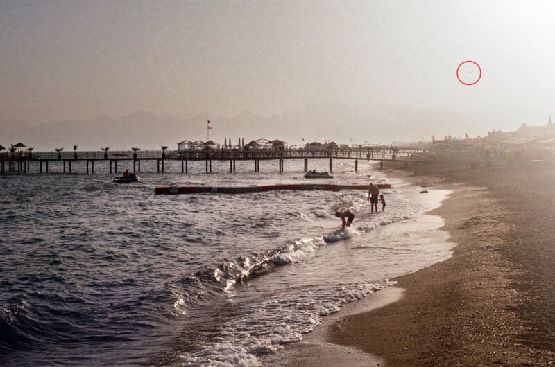 A sandy beach with gentle waves, people walking near the shore and a wooden jetty running out into the sea.  The sun is setting, creating a hazy, warm light.  In the background, mountains are faintly visible through the fog, and a red circle is marked in the sky.