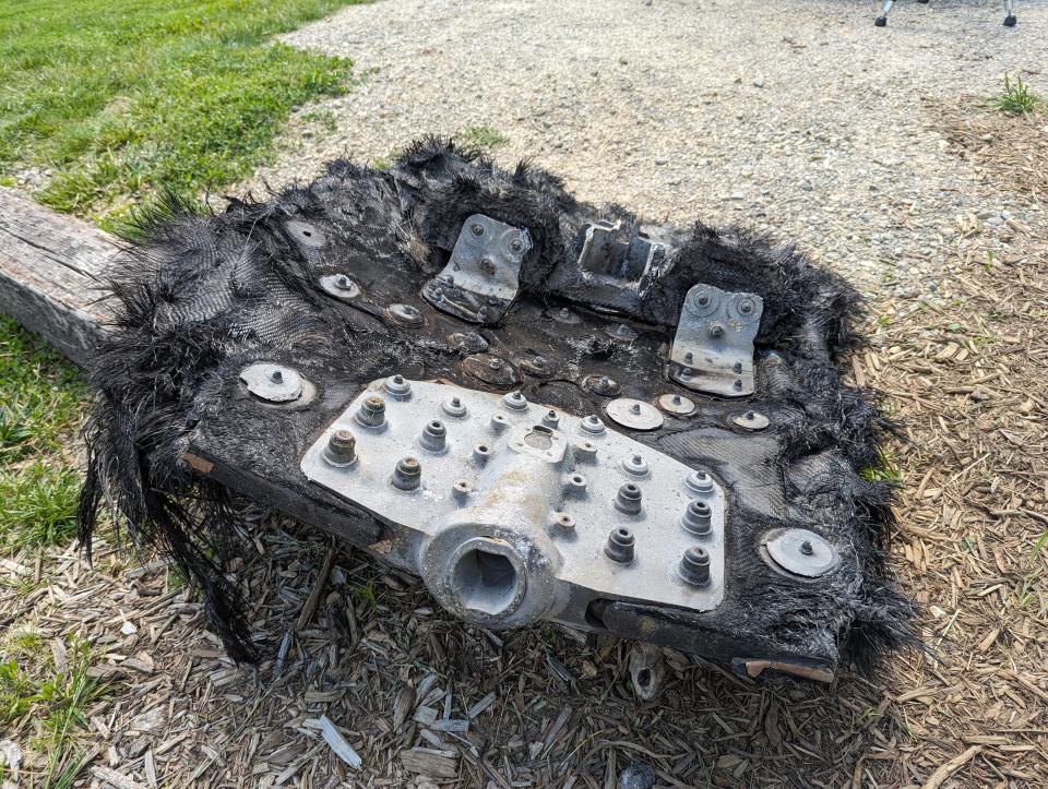 a large black piece of fiberglass covered in metal screws and plates lies on the ground next to a path leading into the forest.  rolling mountains can be seen in the distance