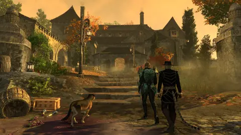 Bethesda's Elder Scrolls Online screenshot shows two humanoid figures – one human and one with a tail – approaching a large mansion-style building at sunset.  A mist of some kind lines the approach, which is littered with barrels and similar objects, and a large feline companion walks beside them.