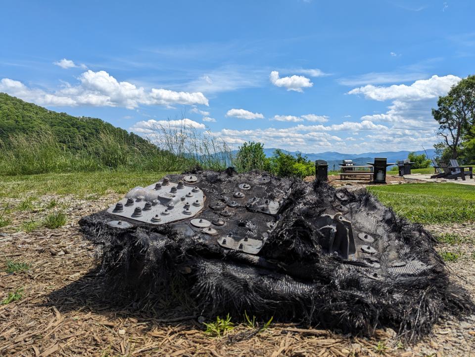 a large black piece of fiberglass covered in metal screws and plates lies on the ground next to a path leading into the forest.  rolling mountains can be seen in the distance