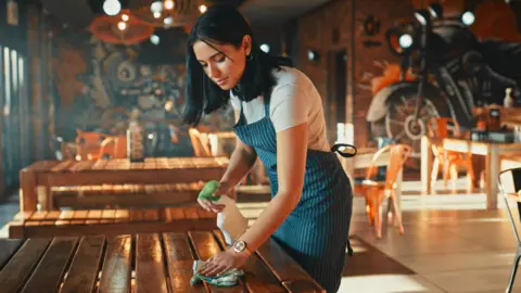 Getty Images A young woman with dark hair works in a cafe and cleans the table