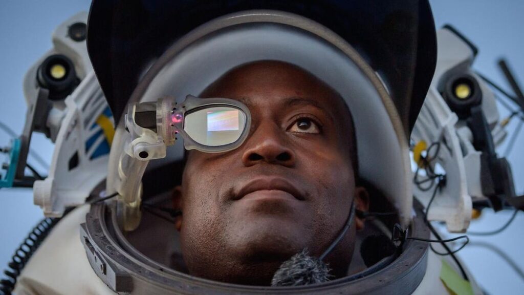 an astronaut looking at the sky in a simulated spacesuit helmet. on his right eye is a small screen used for augmented reality work