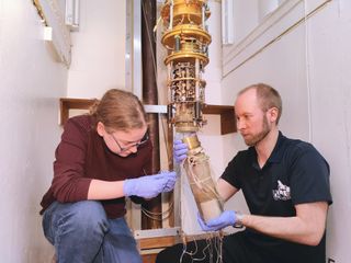 A white room containing two crouched people tending to an intricate golden machine