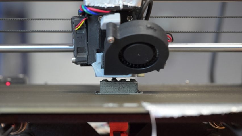 Close-up photo of a 3D printer producing a small gray Lego-like block