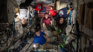 several astronauts in a dark space station module with their faces and bodies illuminated by camera lighting.  there is an american flag in the back