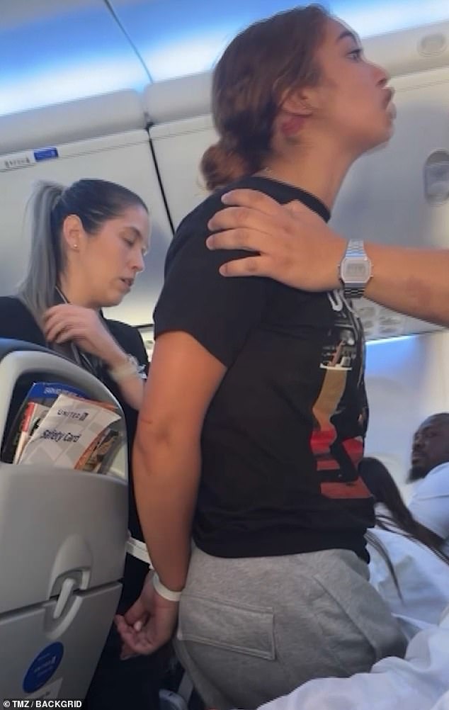 The video cuts to the woman, who now has her hands zipped behind her back and is standing in the aisle yelling at everyone not to fly United