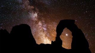 the rock formation in the foreground is shaped like an arch, while the star-studded sky in the background shows the band of the Milky Way stretching across the sky.