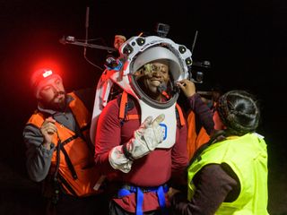 the astronaut in the center of the image in a simulated helmet and space suit.  two engineers on each side adjust his helmet