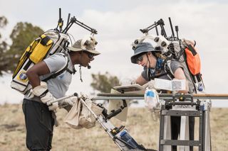 two astronauts leaning over a table in a field while wearing backpacks with antennas on top