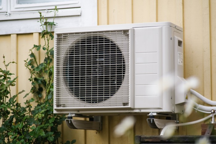 An outdoor air conditioning unit is attached to the wall of the yellow house, showing the fan inside the metal casing