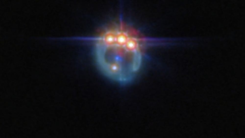 A small image of a galaxy distorted by gravitational lensing into a dim ring. At the top of the ring are three very bright spots with diffraction spikes coming off them, right next to each other: these are copies of a single quasar in the lensed galaxy, duplicated by the gravitational lens. In the centre of the ring, the elliptical galaxy doing the lensing appears as a small blue dot. Courtesy: ESA/Webb, NASA & CSA, A. Nierenberg